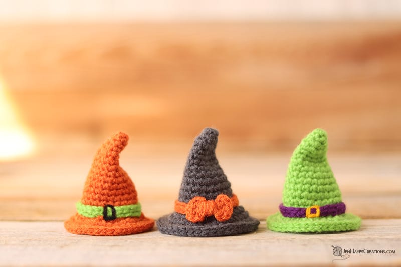 Three small crochet witch hats in orange with a green band, grey with an orange bow, and green with a purple band sitting on a wooden surface.